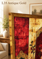 a close-up view of a gold-colored antique frame with a red and gold floral design, placed on a wooden surface. In the background, there is a window with a plant and a painting on the wall.